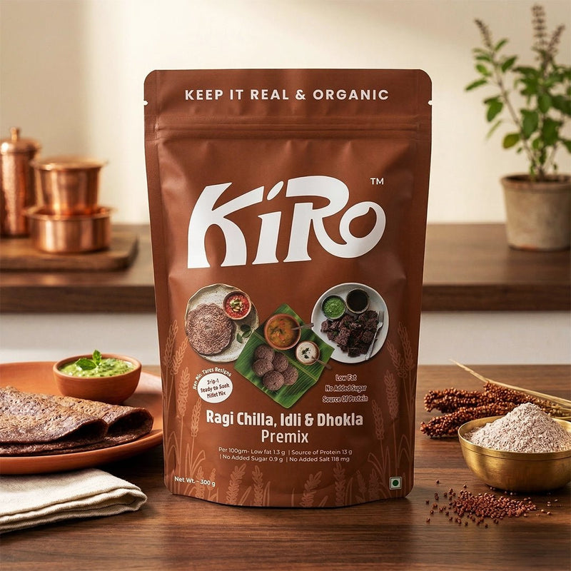 Kiro Ragi Chilla Idli Dhokla 3-in-1 premix 300g pouch on wooden table with copper kitchenware and finger millet grains